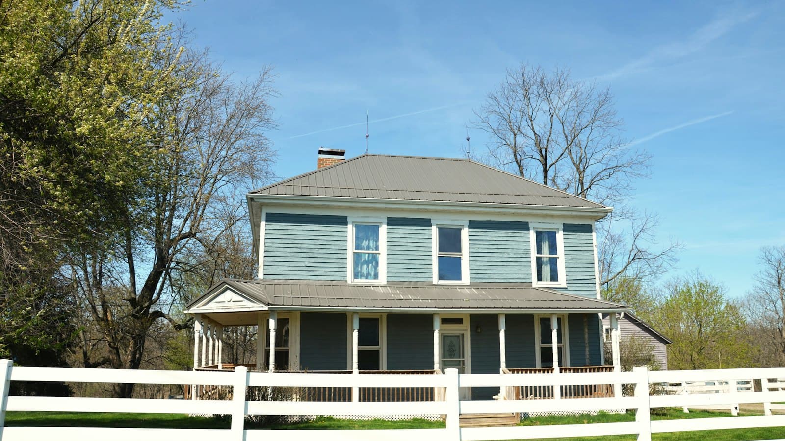 Stock concept image of a two-storey farmhouse with a wraparound porch behind a white rail fence — not an actual Cali Craft Renovations project